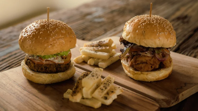 Two gourmet burgers with sesame seed buns on a wooden serving board, with grilled cheese sticks in the background.