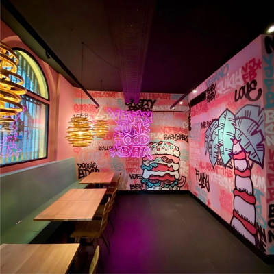 Colorful graffiti-style mural inside a themed restaurant or cafe, featuring a large bubblegum with lips, a candy cane, and various words and phrases in vibrant neon colors. Wooden tables and chairs are along the left side.