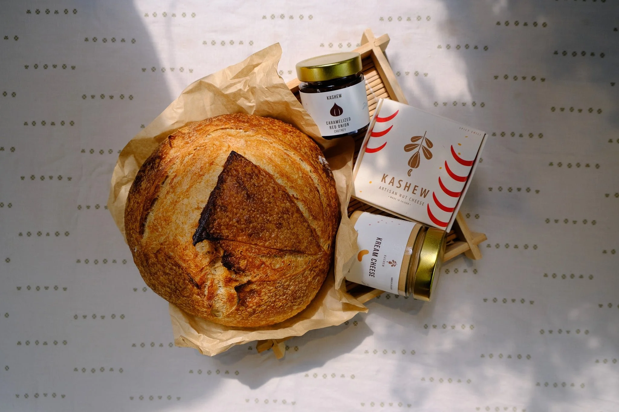 A round, rustic loaf of bread with a golden-brown crust on crumpled parchment paper, surrounded by jars of flavored cheese spreads and a box labeled 'Kashew Artisanal Nut Cheese' on a light-colored surface with small, gold decorative markings.