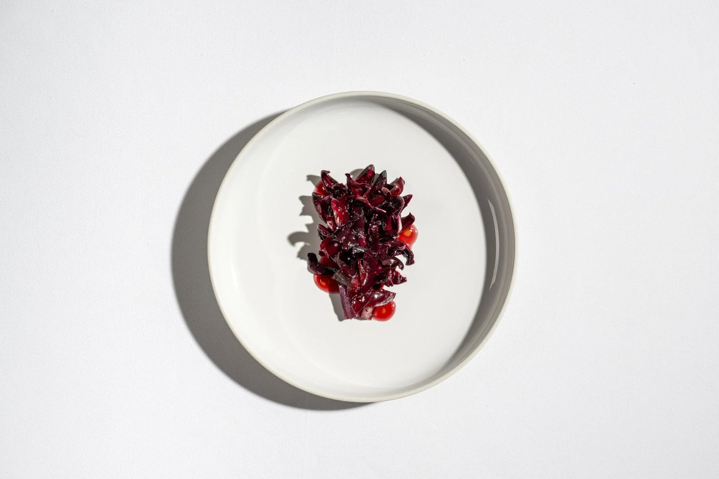 A white plate with a serving of leafy greens, possibly beet greens or red kale, garnished with small red berries or pomegranate seeds, on a white surface.