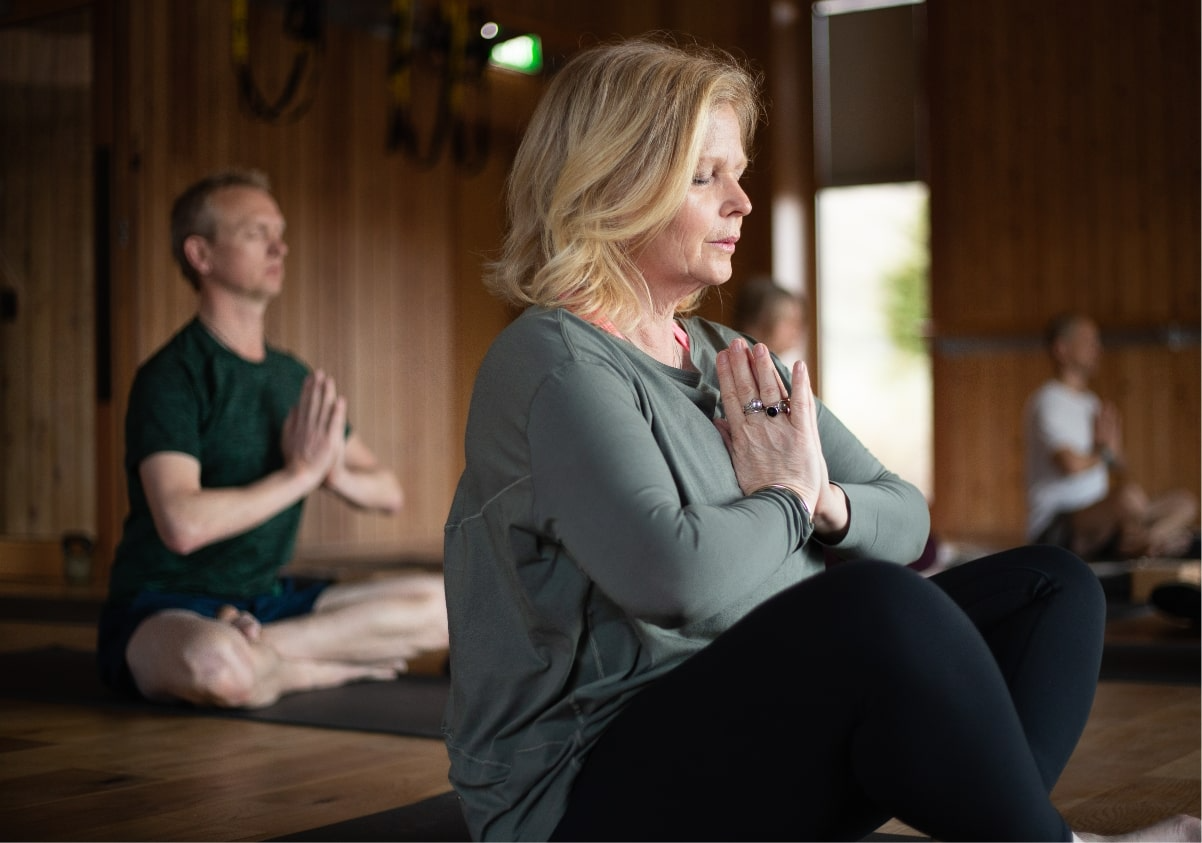 People practicing yoga inside a wooden studio, sitting cross-legged with hands in prayer position, eyes closed, focusing inward.