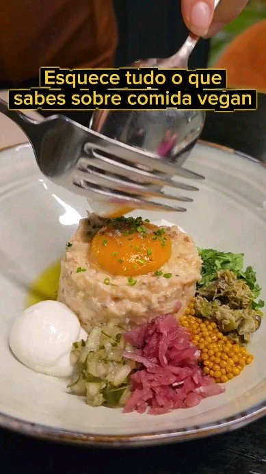 A plate of vegan food with a textured patty topped with a fried egg, surrounded by a dollop of white sauce, green relish, pink pickled vegetables, mustard seeds, and chopped herbs. A person is holding a fork and spoon over the dish, and there is a ca