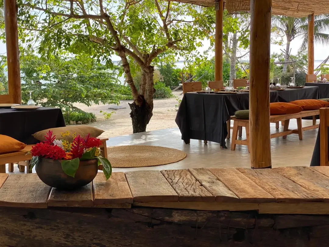 Outdoor dining area with wooden tables and benches, black tablecloths, glassware, and pillows, overlooking a sandy beach with trees and ocean in the background.