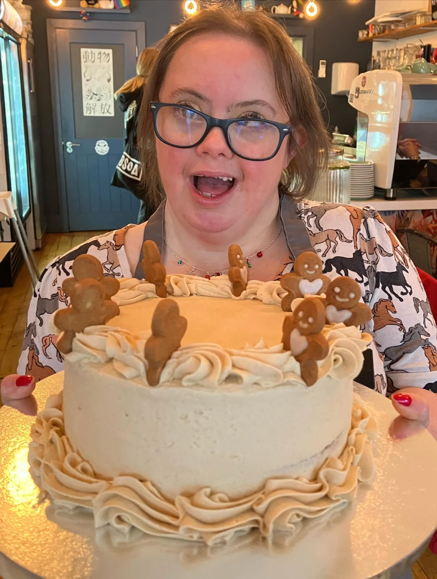 A woman with glasses and a patterned shirt is holding a white birthday cake decorated with gingerbread cookies and cream frosting. She is smiling with an open mouth in a restaurant or cafe setting.