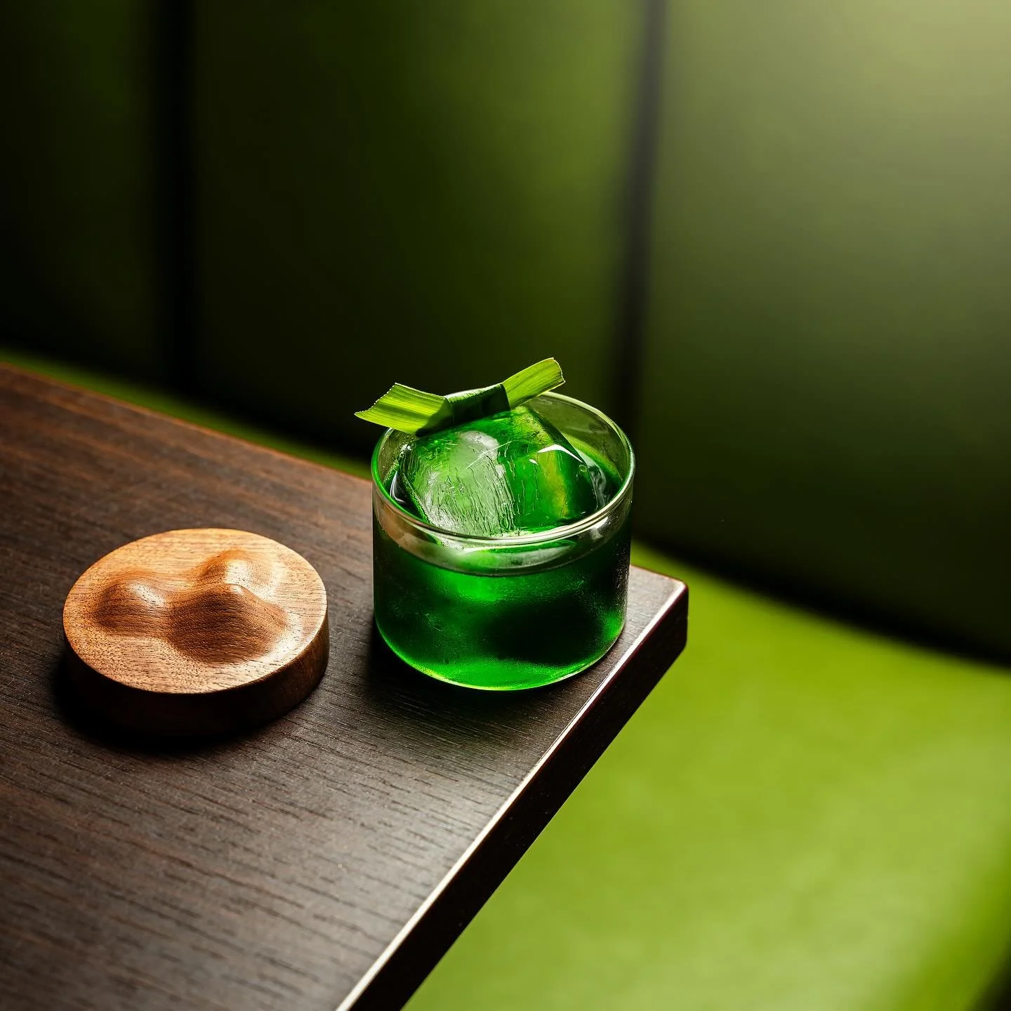 Green drink with ice cubes and a leafy garnish in a clear glass on a wooden surface, with a small wooden dish nearby.