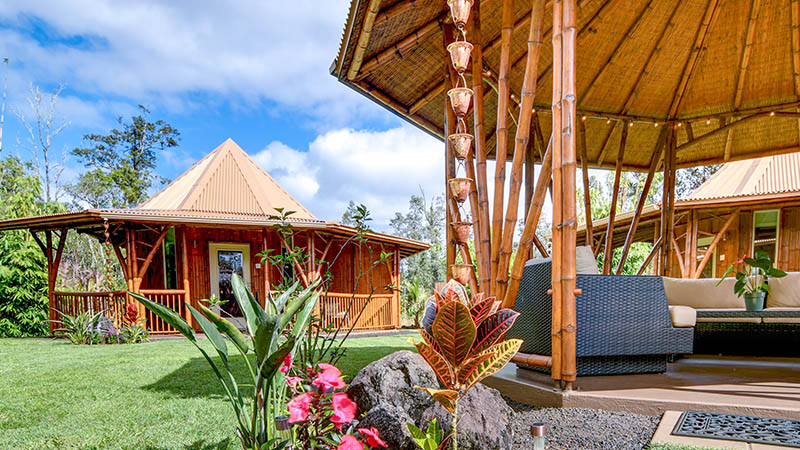 Outdoor view of bamboo and wooden cabin with patio furniture, lush green lawn, colorful flowers, and trees under a bright blue sky.