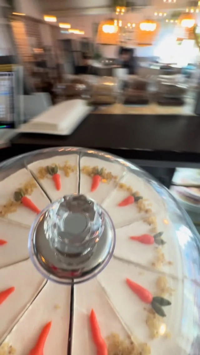 A pizza with a clear plastic cover, topped with small carrot and green pepper decorations, on a table near a busy restaurant counter with blurred background.