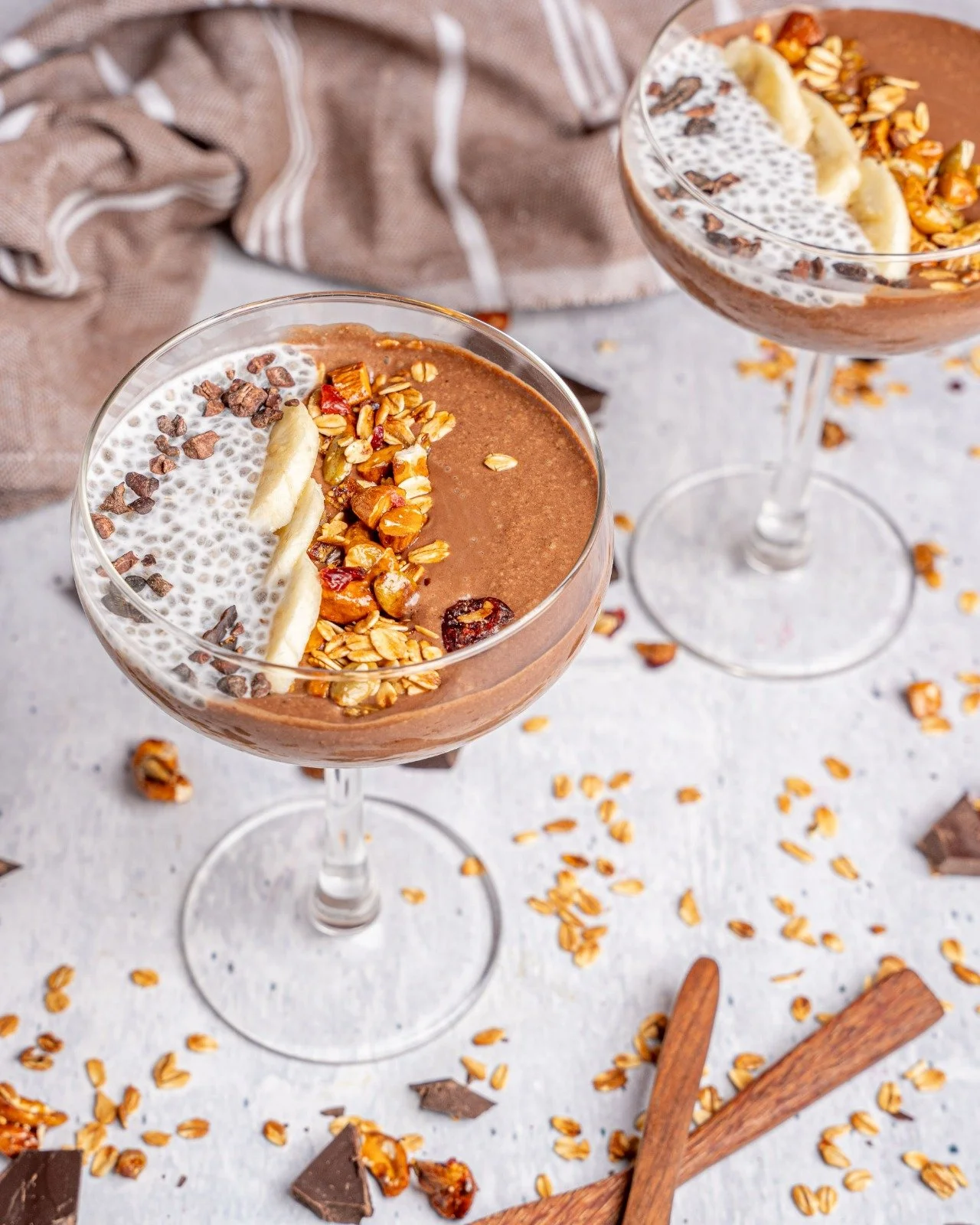 Two glass dessert cups filled with chocolate mousse topped with sliced banana, granola, cocoa nibs, and shredded coconut, placed on a white surface with scattered oats, chocolate pieces, and cinnamon sticks around.