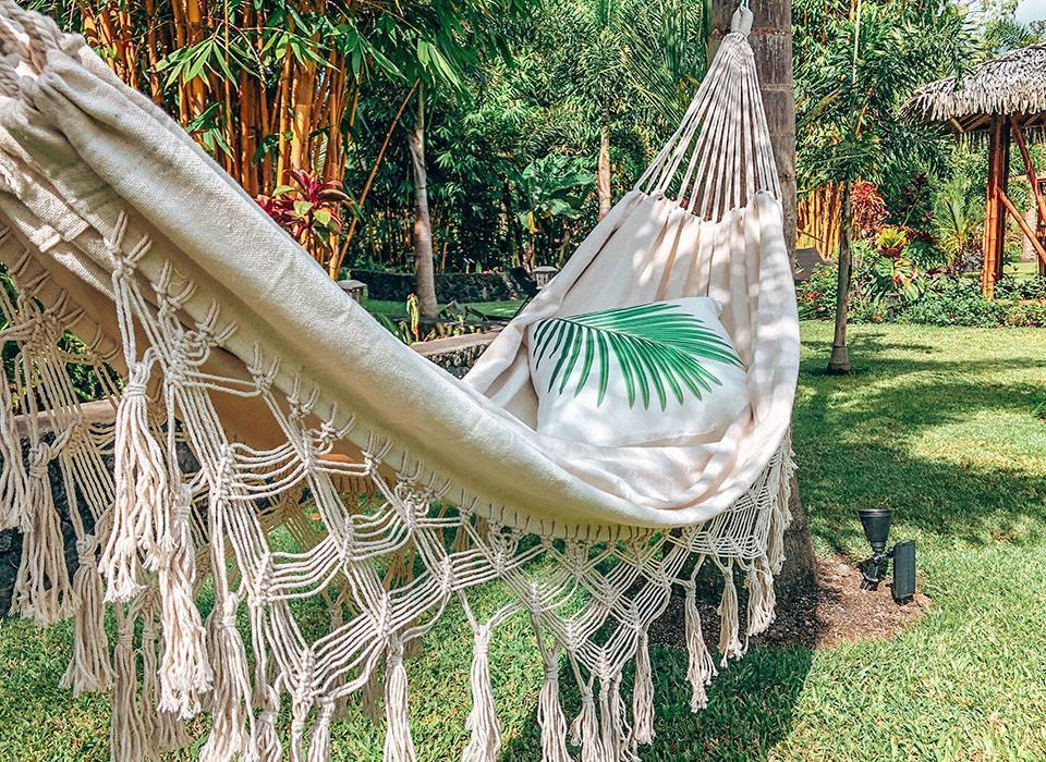 A white hammock with fringed edges hanging between trees in a lush, tropical garden with green plants and a thatched hut in the background. A pillow with a green palm leaf print is inside the hammock.