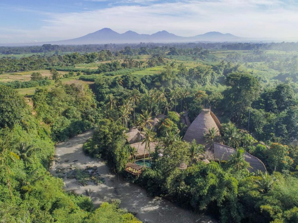 Aerial view of lush green tropical forest with a river flowing through it, traditional thatched-roof structures partially hidden among trees, and mountains in the distance.