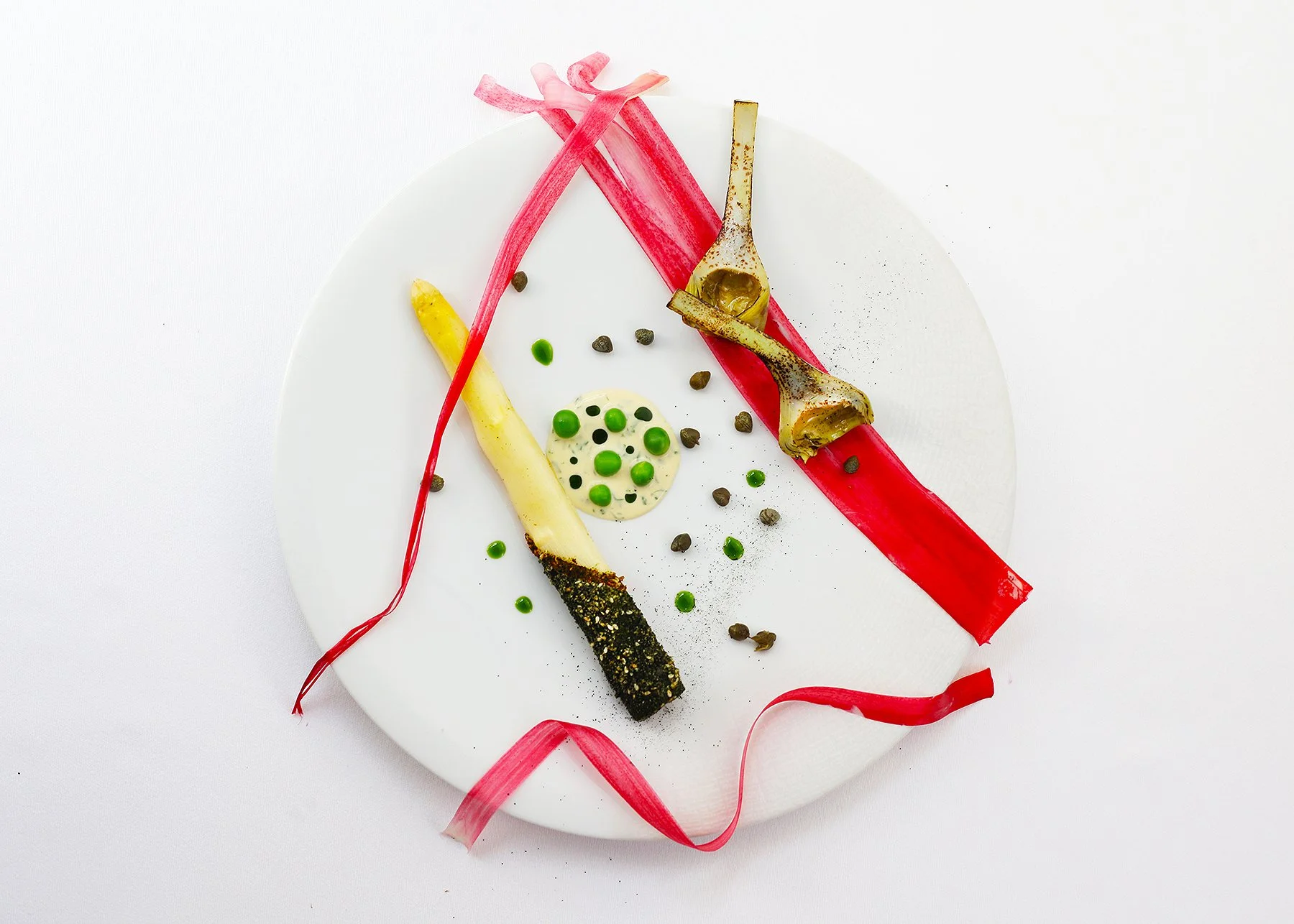 Artfully plated gourmet dish on a white round plate featuring roasted vegetables, green peas, and a creamy sauce, garnished with a pink ribbon and black pepper.