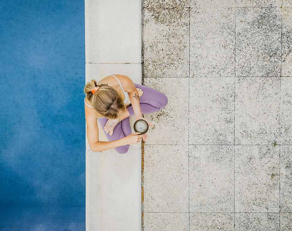 A woman practicing yoga outdoors, sitting cross-legged on patterned tiles, holding a small bowl, with a blue mat beside her.