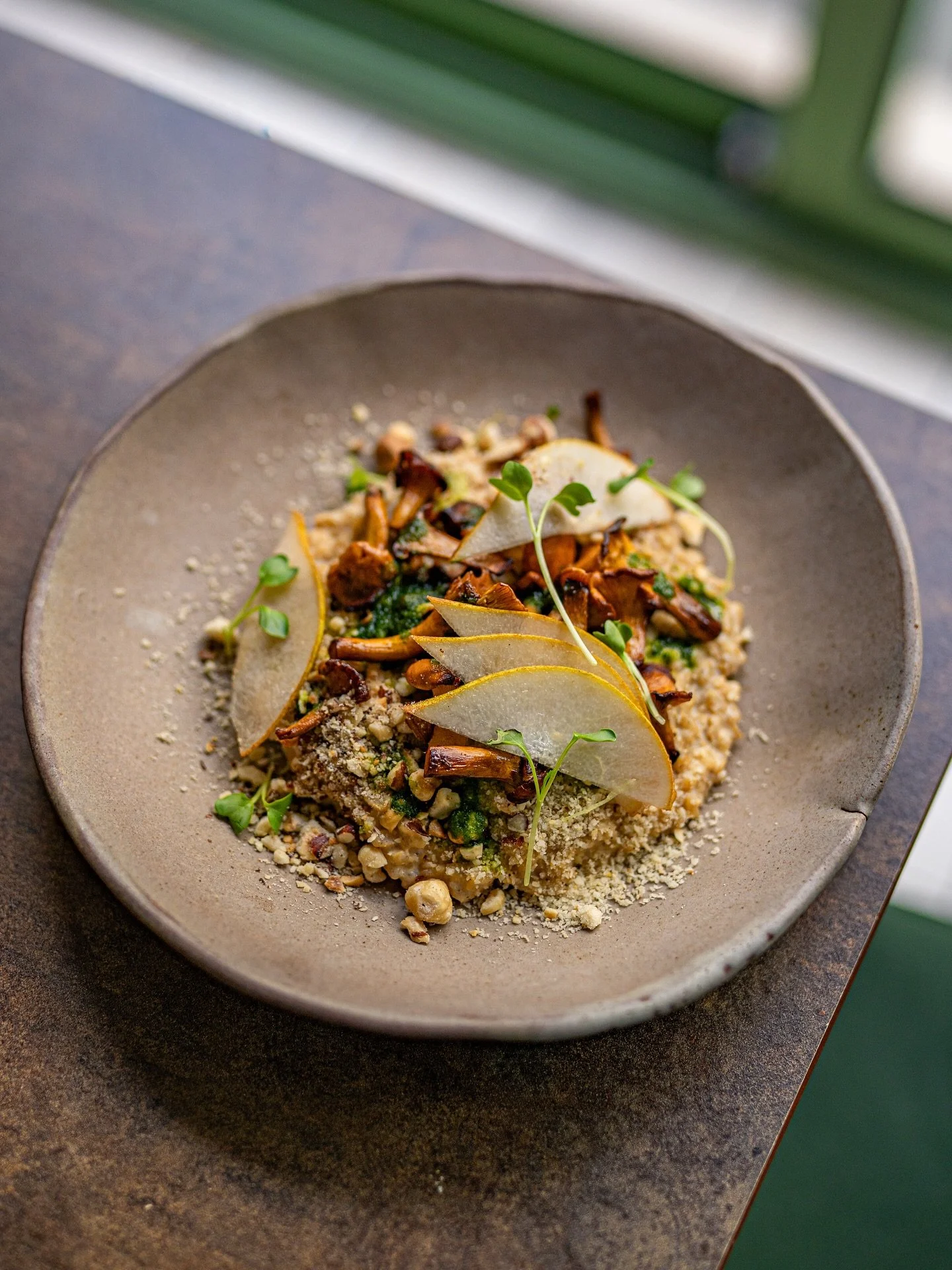 A plated dish with various garnishes on a ceramic plate, including sliced pears, microgreens, crispy crunchy elements, and small chopped greens, set on a dark brown table near a window with green window frames.