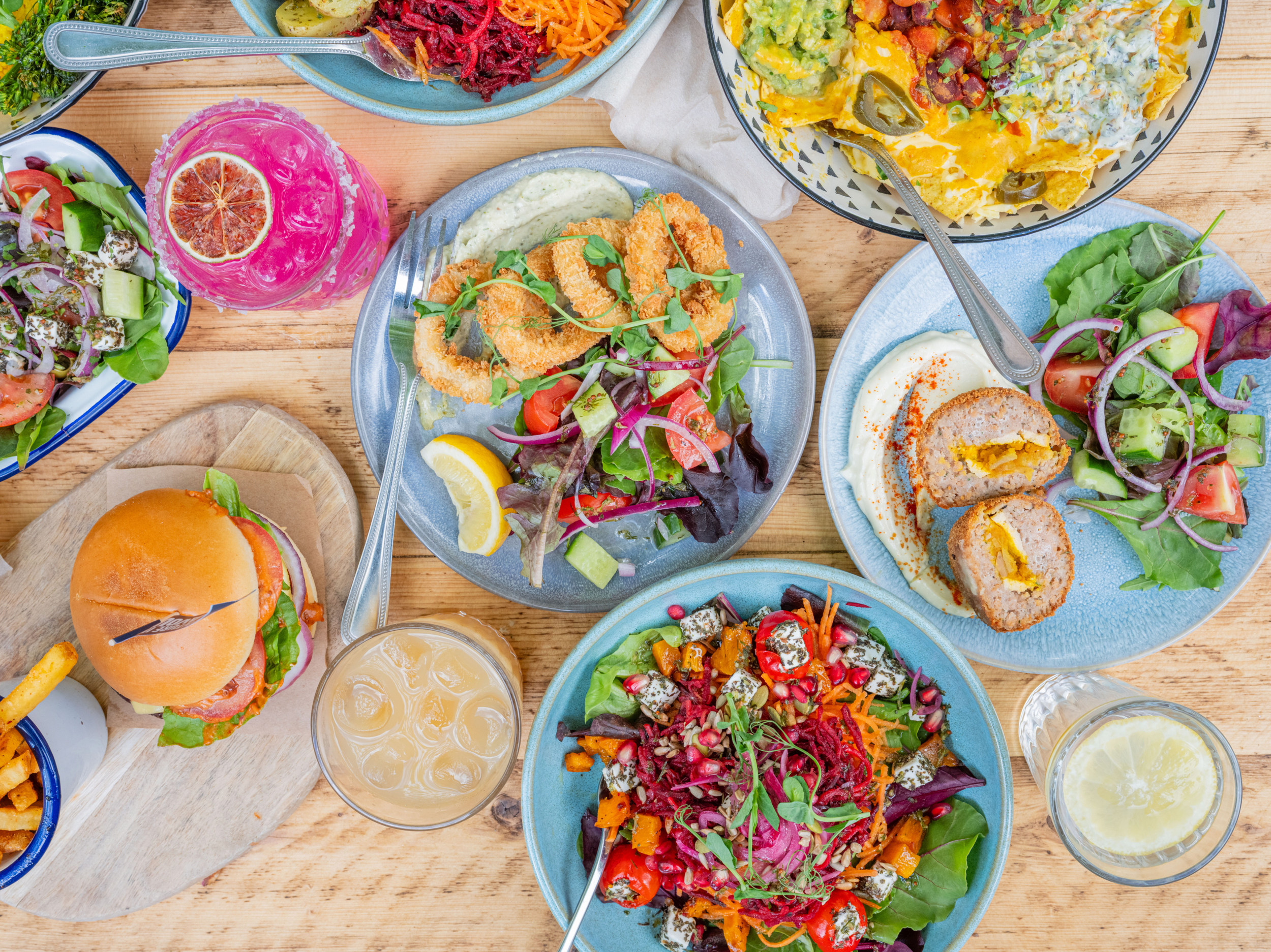 Assorted dishes on a wooden table, including salads, a burger, a pink-colored beverage with lemon, a plate of fried fish with sauce, and various other colorful foods.