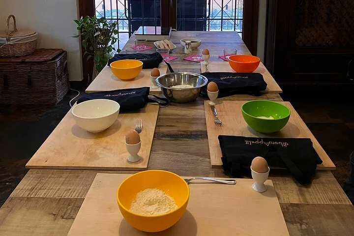 A kitchen table set for a cooking or baking activity. The table has multiple bowls, some of which are yellow, orange, green, and white, with eggs in egg cups beside some bowls. There are also utensils, black napkins, and a large mixing bowl in the ce
