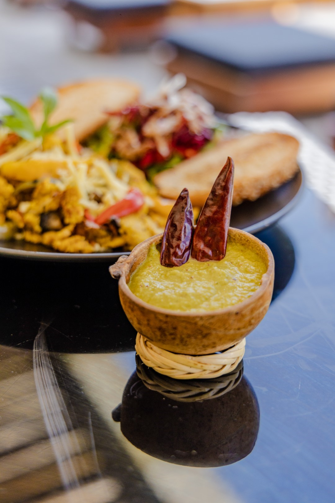 A bowl of green guacamole topped with two dried red chili peppers, placed on a woven coaster on a reflective black table, with plates of food in the background.