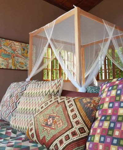 Canopy bed with sheer curtains, multiple patterned pillows, and a colorful quilted bedspread in a room with large windows and a wooden frame.