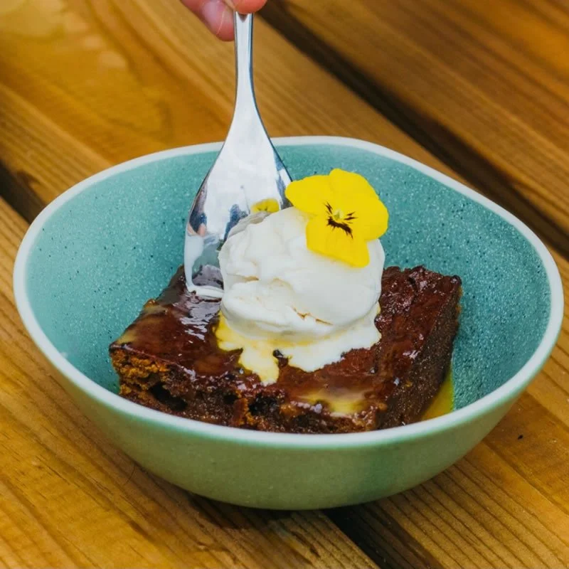 A bowl of brownie topped with vanilla ice cream and a yellow edible flower, on a wooden table.