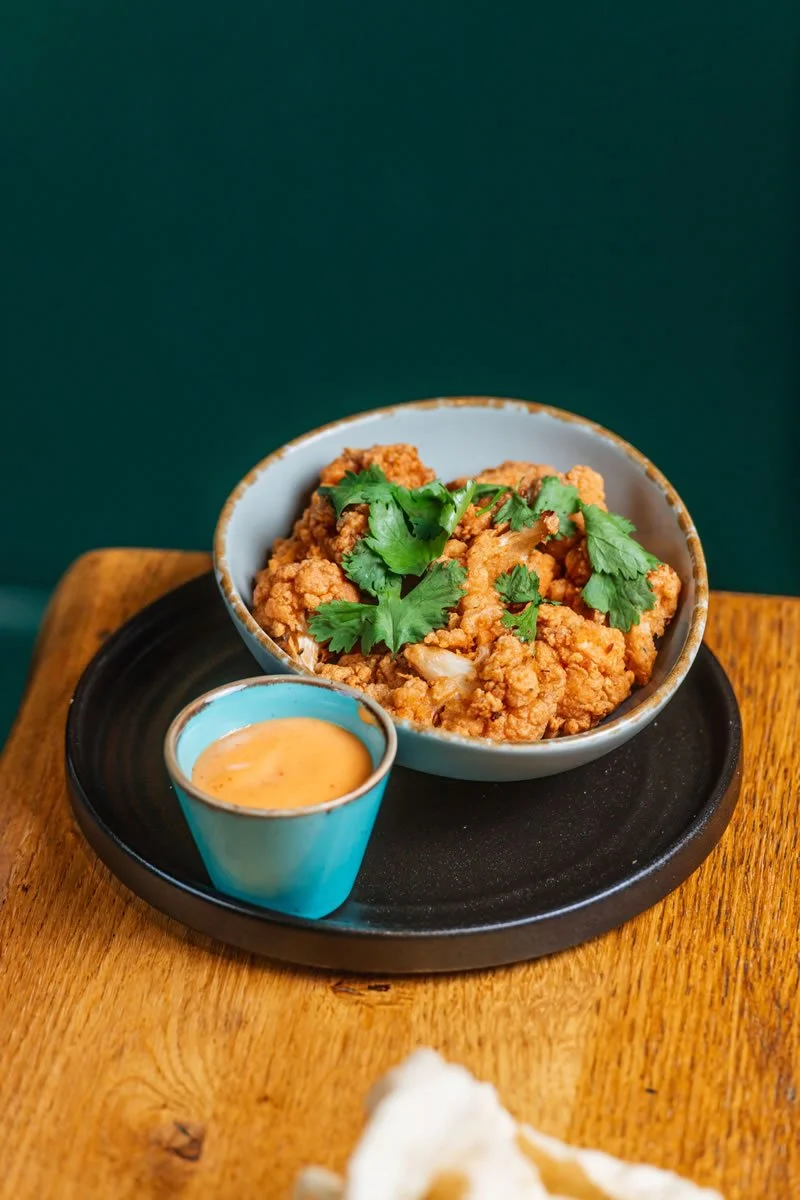 Bowl of fried chicken garnished with cilantro on a black tray with a small cup of dipping sauce on a wooden table.