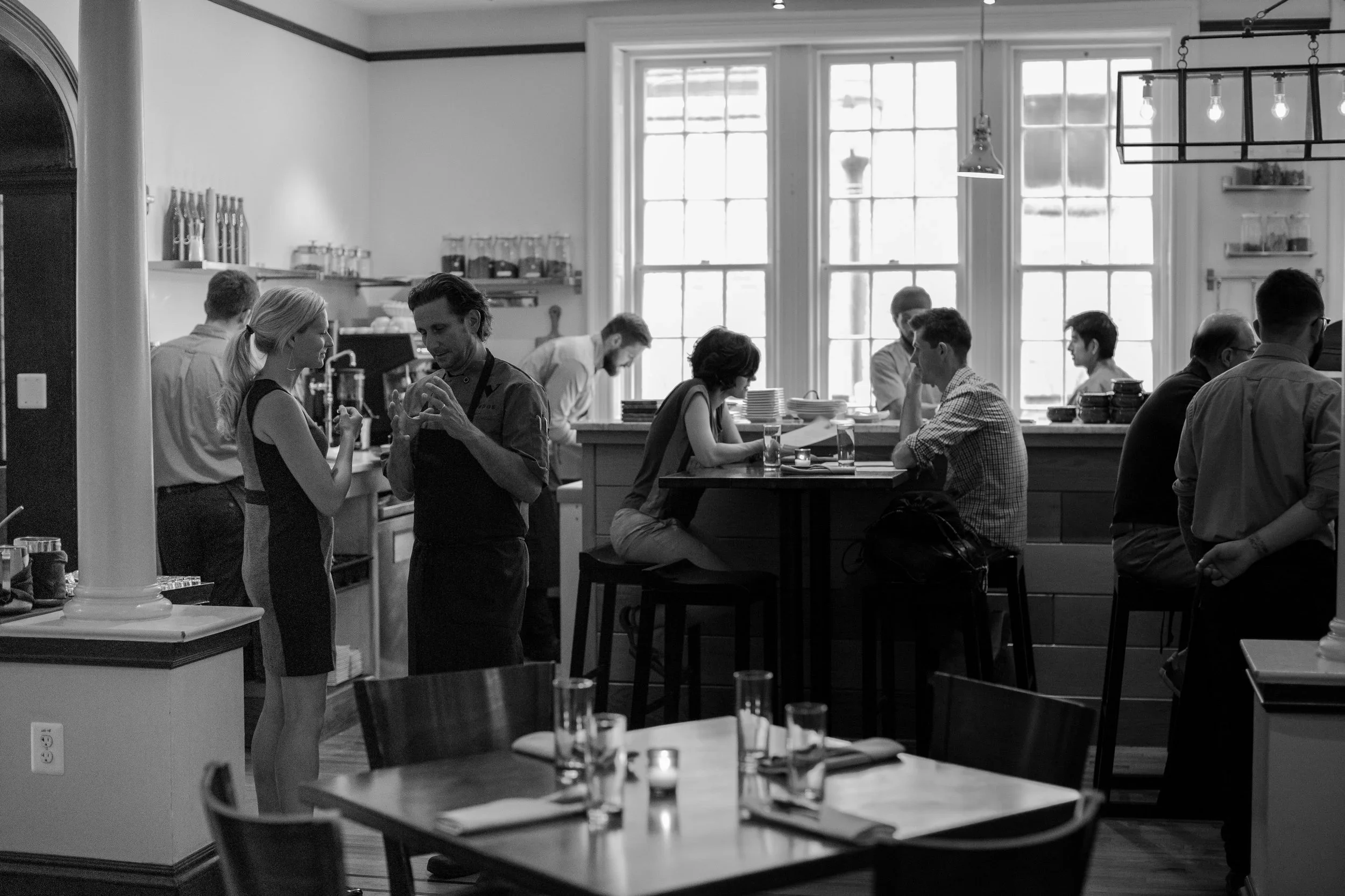 A busy restaurant or café with several patrons and staff. Two women are engaged in conversation near the front, while others are seated at tables or working behind the counter. The interior features large windows, modern lighting, and shelves with ja