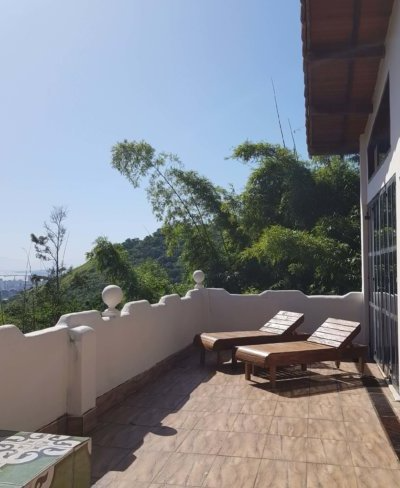 Outdoor balcony with two wooden lounge chairs, white railing, tiled floor, and lush green trees in the background under a clear blue sky.