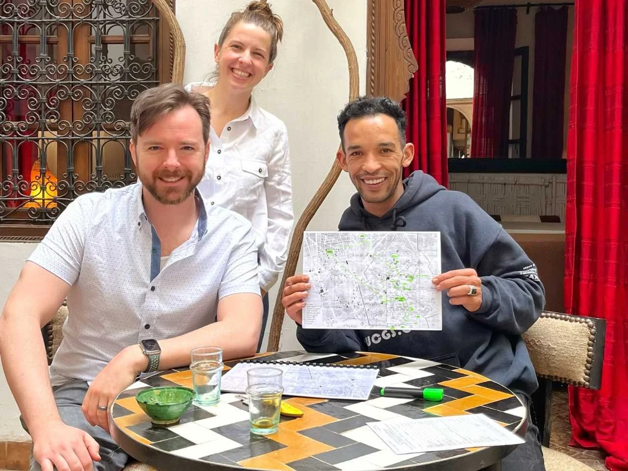 Three people sitting around a table, two men and one woman, smiling. The man on the right is holding a map, and there are drinks on the table. Red curtains and ornate woodwork are in the background.