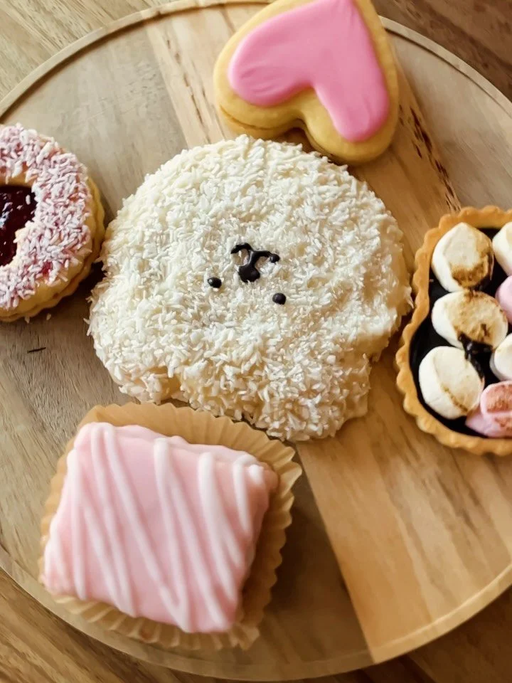 Assorted decorated cookies on a wooden platter, including a bear-shaped cookie with white coconut flakes and black icing, a pink heart-shaped cookie, a small marshmallow-topped tart with chocolate and marshmallows, a square pink frosted cookie, and a