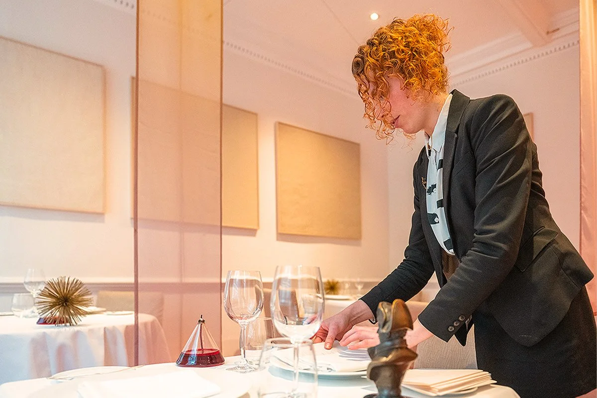 A woman setting a table in an elegantly decorated restaurant with glassware, plates, and decorative items on the table.
