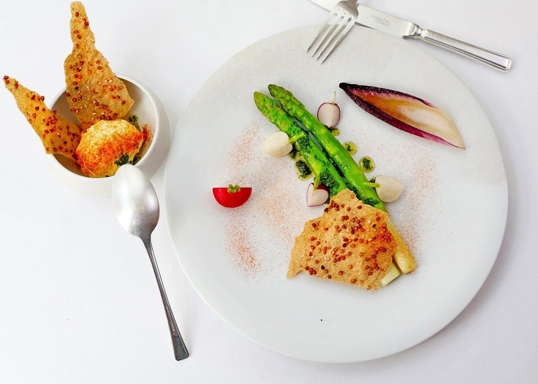 Plated gourmet dish with green asparagus, radishes, a purple okra pod, and crispy chips on a white plate. A small bowl with additional crispy chips is beside the plate. Silver fork and knife rest on the top right of the plate on a white background.