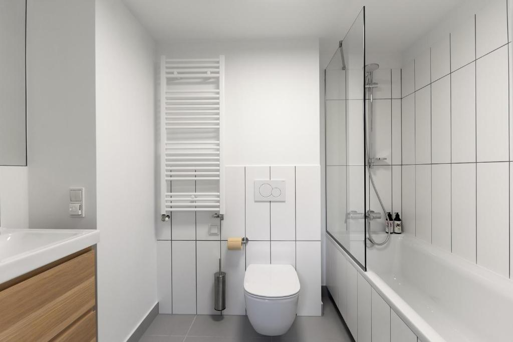 Modern bathroom with white tiles, a wall-mounted toilet, a bathtub with a glass shower partition, a white towel radiator, a toilet paper holder, and a small vanity with a wooden drawer.
