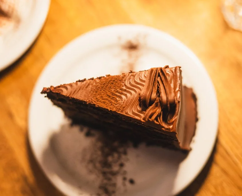 Slice of chocolate cake on a white plate with chocolate frosting and decoration, on a wooden table.