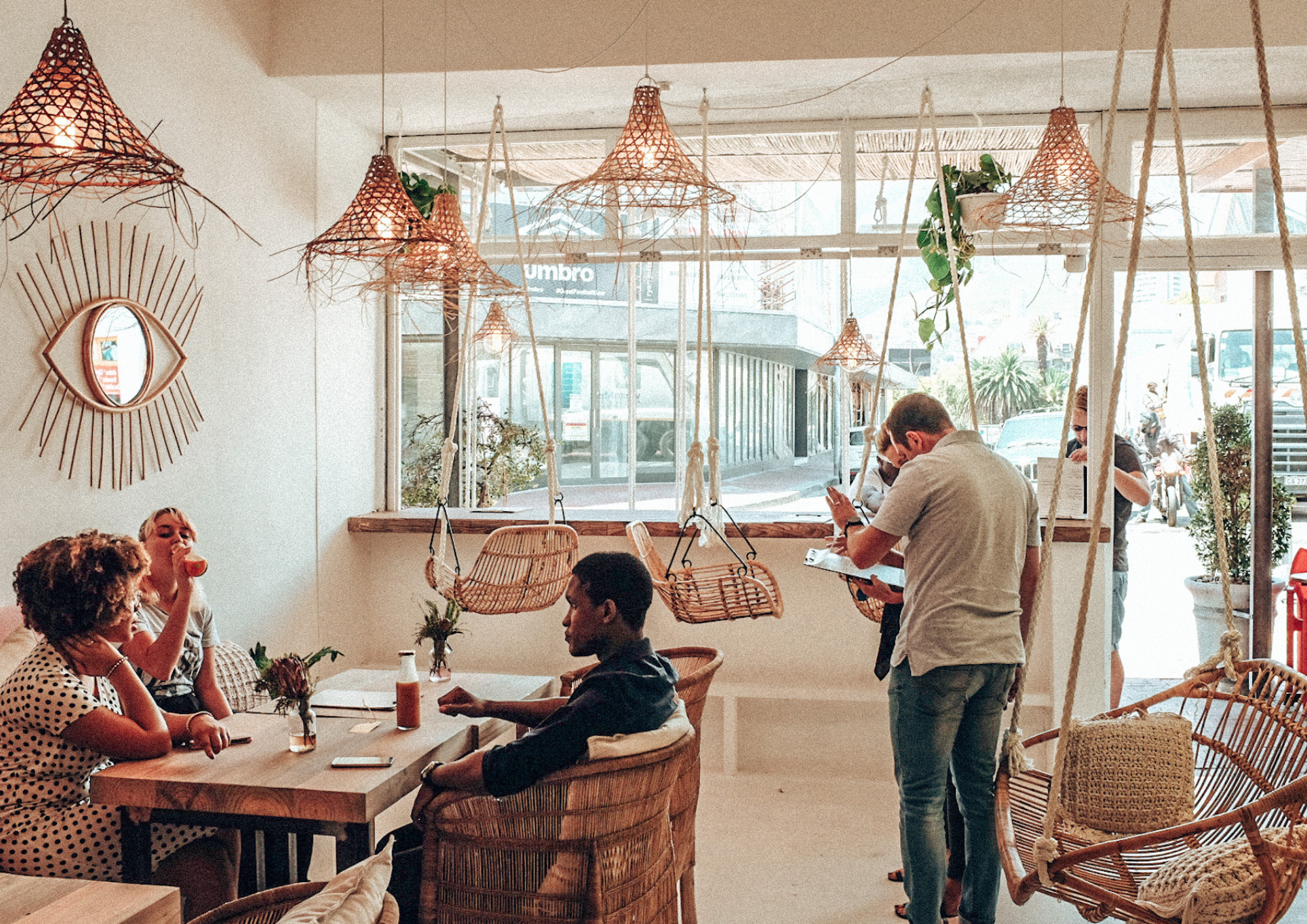 People dining inside a cafe with hanging wicker lamps, wooden and wicker furniture, and large windows showing the street outside.