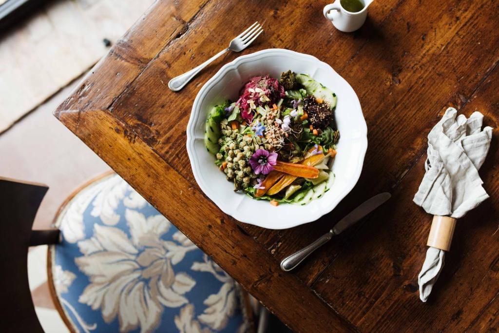A bowl of colorful vegetable salad on a wooden table with cutlery, a napkin, and a small pitcher in the background.