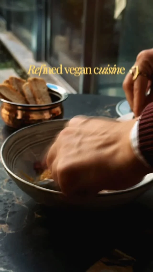 Person placing food into a bowl at a restaurant with the text 'Re/amed vegan cuisine' on the window