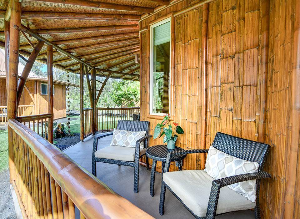 Two black wicker chairs with beige cushions and patterned pillows on a wooden porch, with a small round black table holding a potted plant. The porch has a wooden railing, and the house's exterior wall is made of wooden panels. There is a window with shutters, and trees and grass are visible in the background.