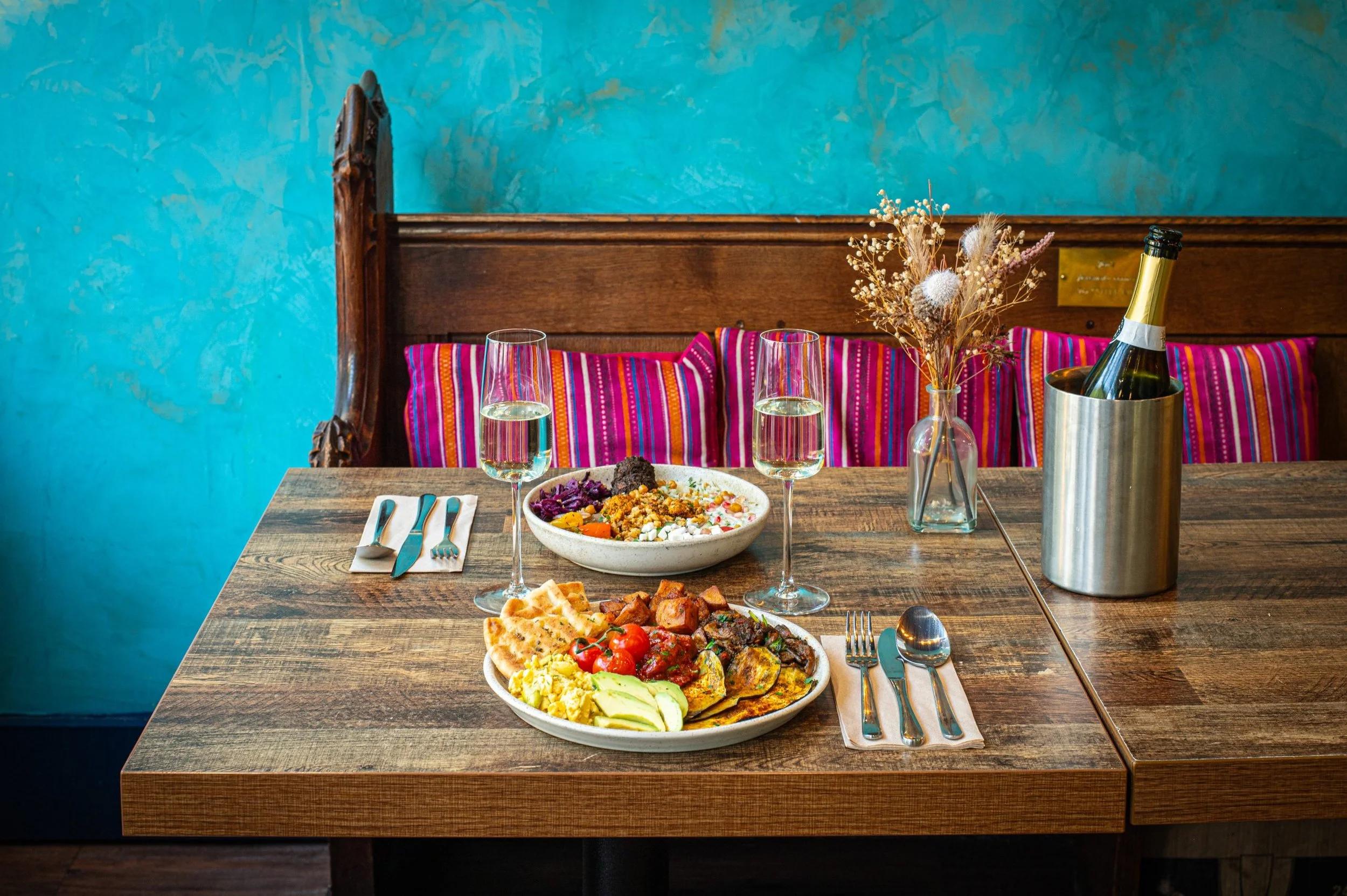 A wooden dining table set with plates of assorted foods, wine glasses, and silverware, with a colorful striped cushion and a flower vase on a bench against a teal textured wall.