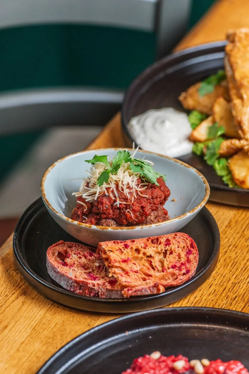 A bowl of chili with shredded cheese and cilantro, served with slices of toasted bread on a black plate, placed on a wooden table.