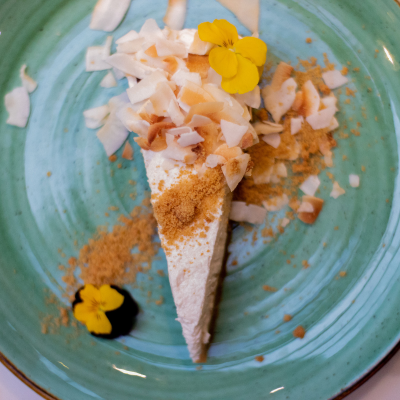 A slice of cheesecake topped with coconut flakes, toasted coconut, and edible yellow flower petals, served on a turquoise plate with coconut shavings and crumbs around it.
