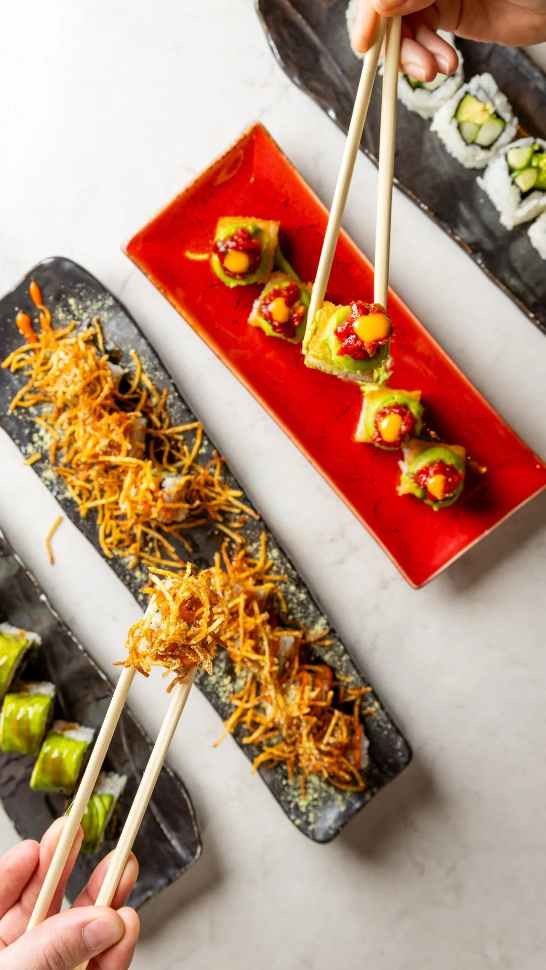 Close-up of sushi rolls and hand holding chopsticks picking up a piece of sushi, with other sushi dishes on black and red plates in the background.