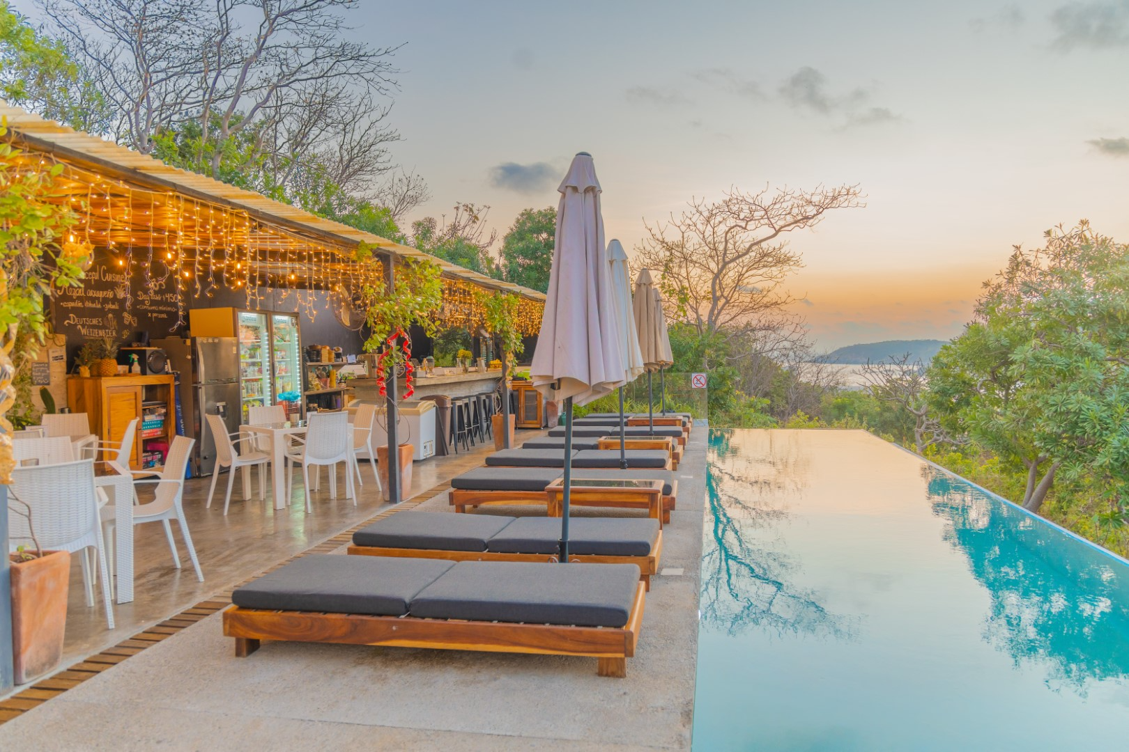 Outdoor poolside bar and lounge area at sunset, with wooden sunbeds, umbrellas, trees, and a scenic view of distant hills and water.