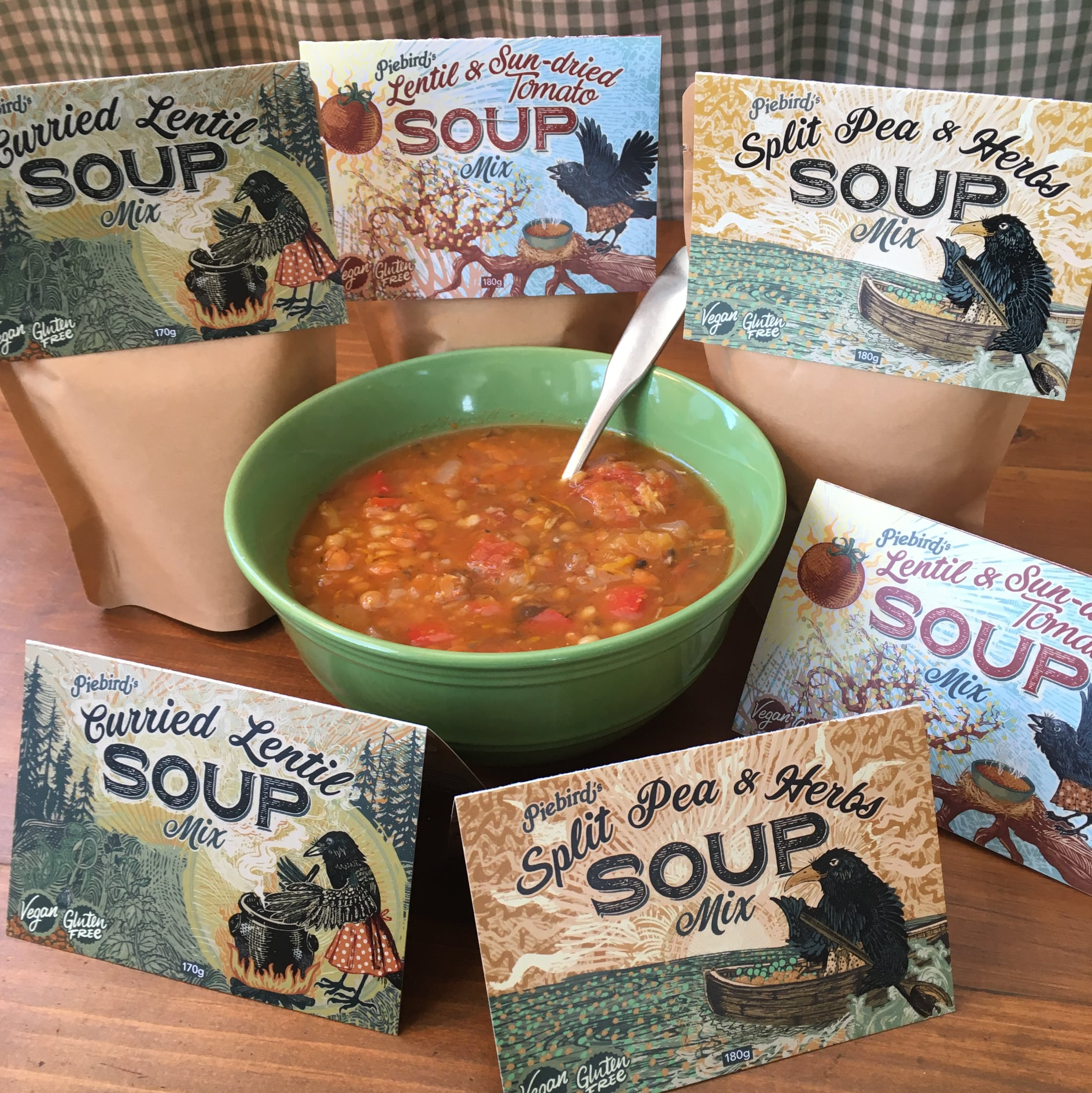 Packages of Piebird's soup mixes, including Lentil & Sun-dried Tomato, Curried Lentil, and Split Pea & Herbs, with colorful illustrated labels featuring birds, trees, and a boat, placed around a green bowl of soup with a spoon.