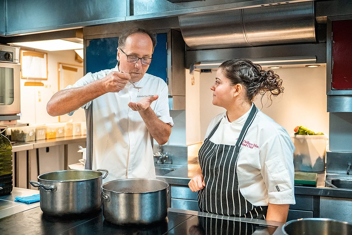 A male chef tasting food in a kitchen, conversing with a female chef who is watching him. Both chefs are wearing white uniforms, and the woman has a black and white striped apron.