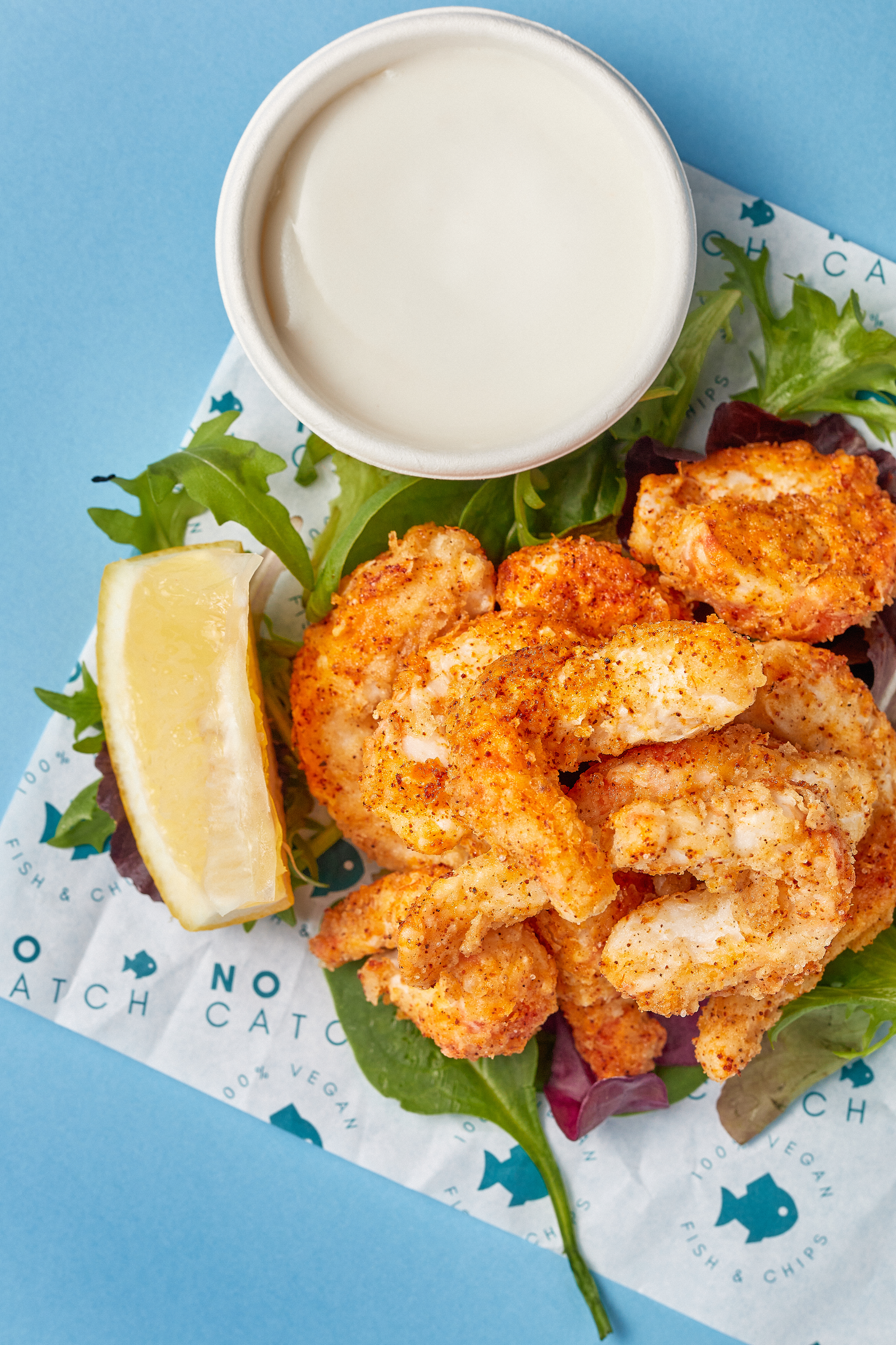 Fried chicken pieces on a bed of mixed greens with a lemon wedge, served with a side of dipping sauce on a blue background