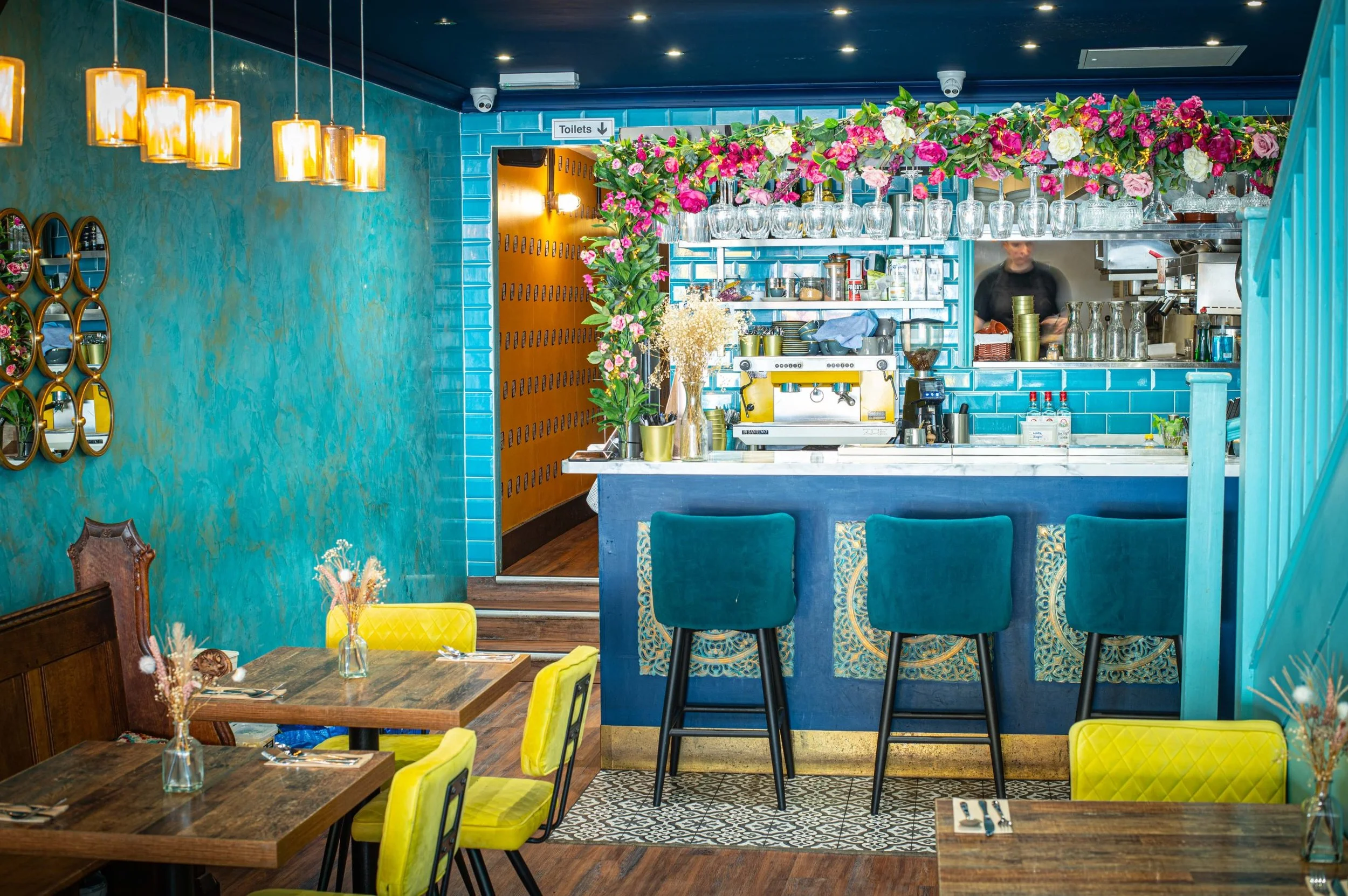 Interior of a colorful restaurant with teal walls, yellow chairs, and a bar area decorated with flowers, glassware, and kitchen equipment.