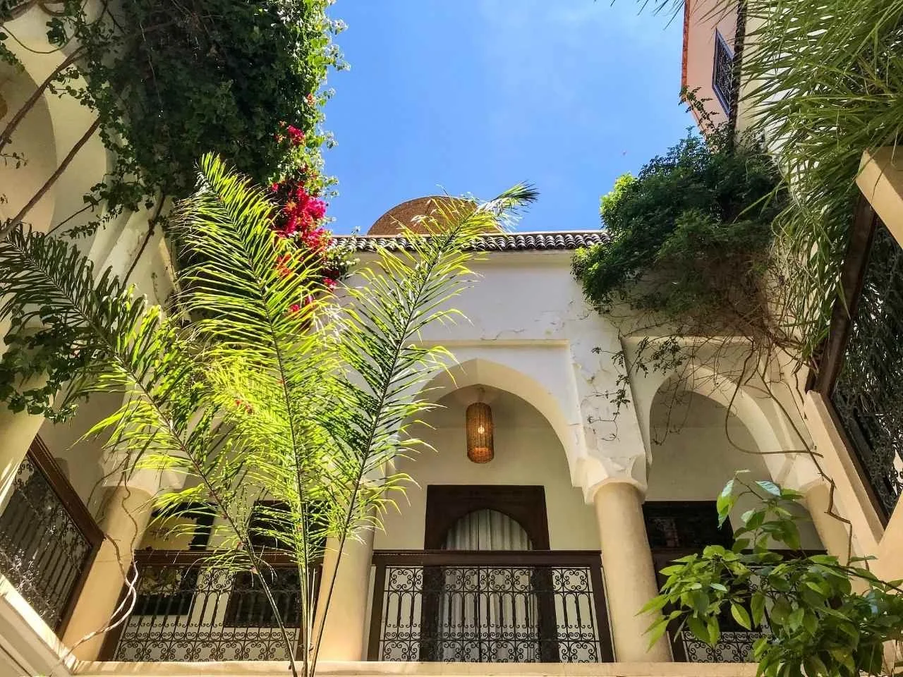 View of a Mediterranean-style courtyard with white stucco walls, black wrought iron balconies, and lush greenery including palm trees and flowering plants, under a clear blue sky.