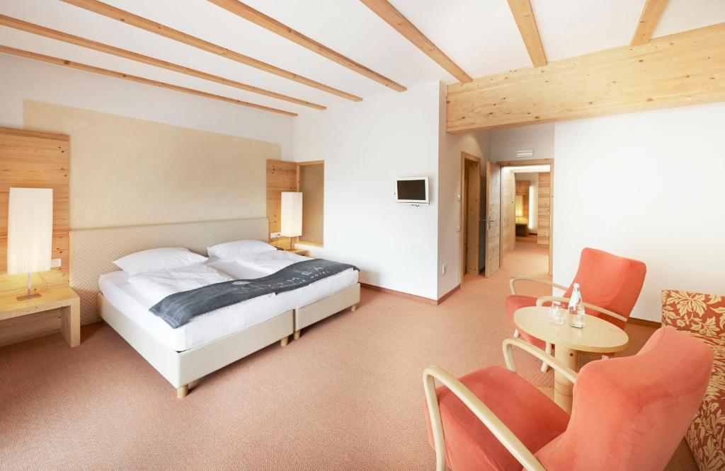 Hotel room with a large bed, side tables with lamps, two pink armchairs, a small round table with water bottles, and a small wall-mounted TV.