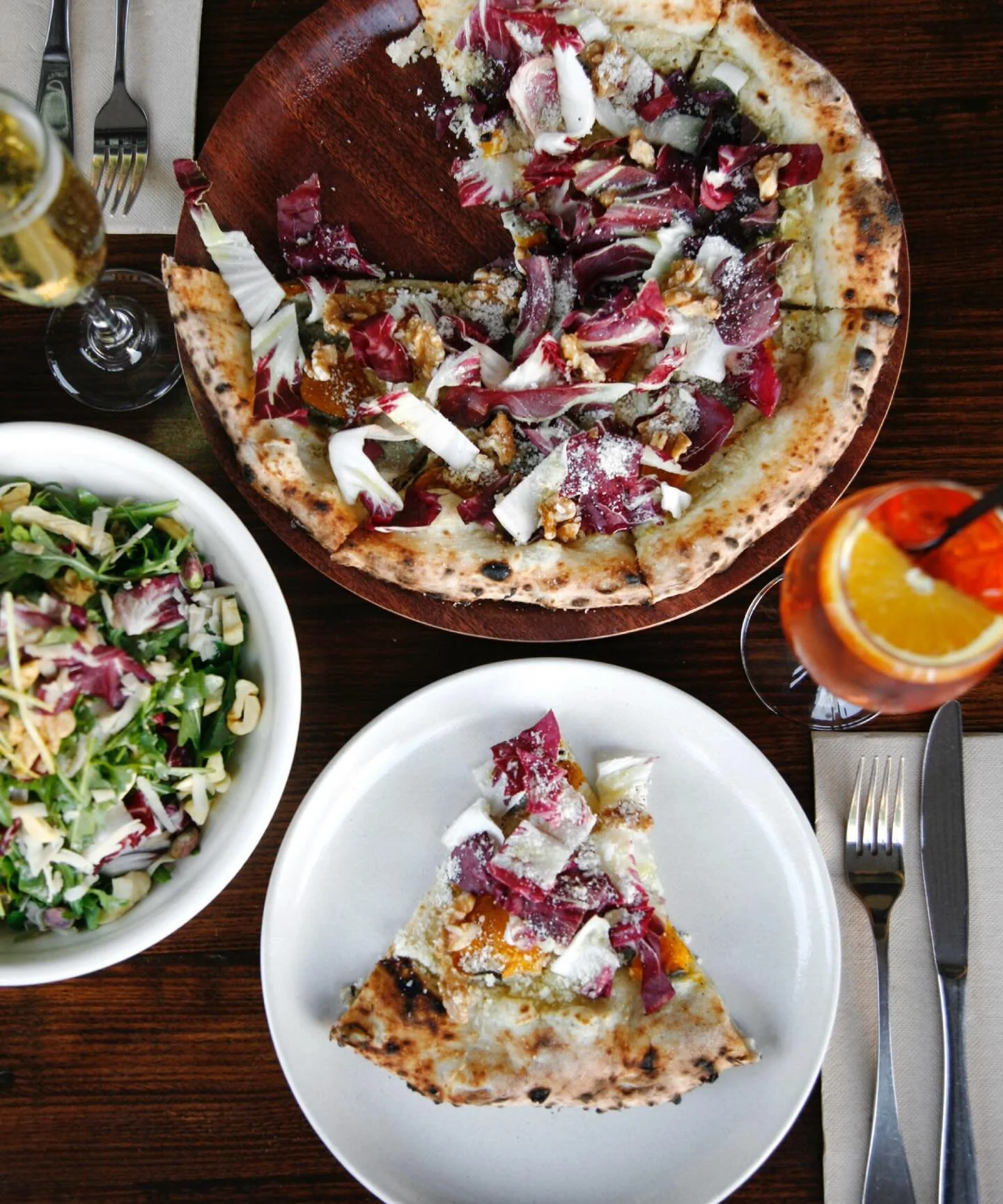 A pizza with toppings including red cabbage, walnuts, and cheese on a wooden board, a salad with greens and walnuts in a white bowl, and a glass of orange-colored drink with ice and orange slices on a dark wooden table.
