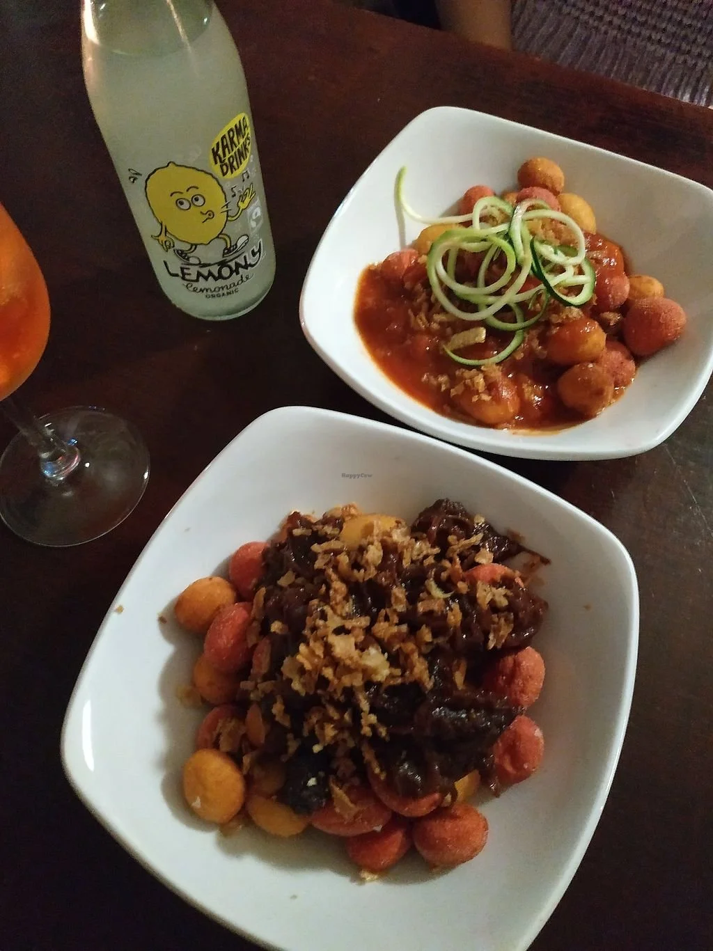 Two white bowls of Asian-style dishes with meatballs and toppings, a bottle of lemon drink labeled "Karma Drinks Lemonade," and a glass of orange wine on a dark wooden table.