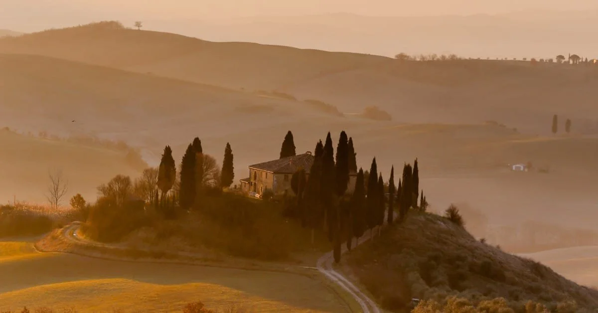 A house on a hill surrounded by tall cypress trees in a daytime landscape of rolling hills and farmland.
