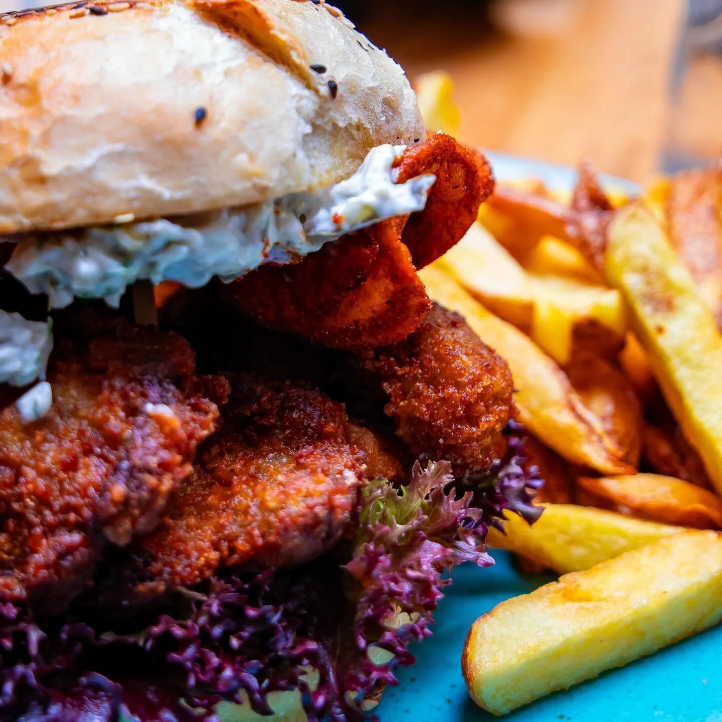 Close-up of a fried chicken sandwich with a bun, fried chicken fillet, lettuce, and mayonnaise, served with French fries and lemon wedges.